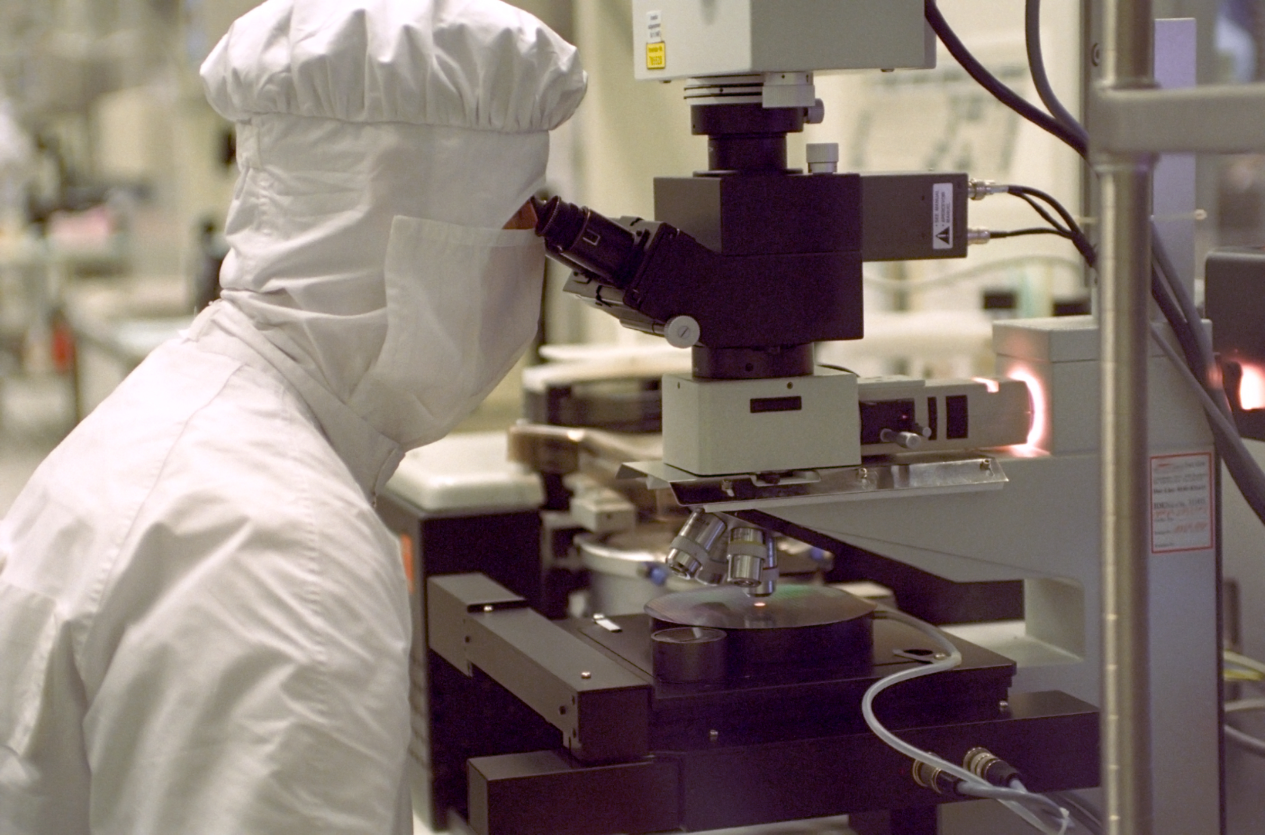 Cleanroom worker in a microchip production plant examining a semiconductor wafer using a microscope.